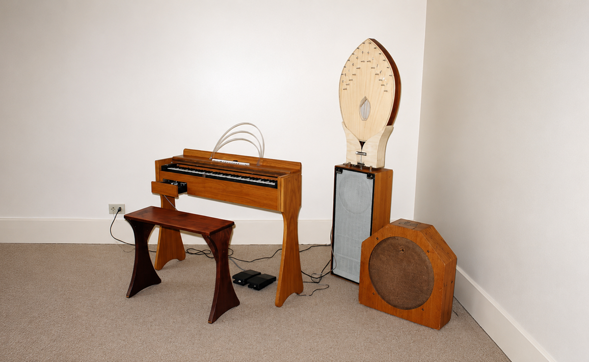 A wooden keyboard instrument with a matching bench sits in the corner of a carpeted room, accompanied by two unusual wooden speaker-like structures, one with an arched top and strings, the other hexagonal with a round front.