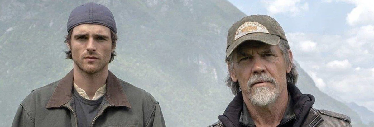 Two men stand outdoors in front of misty mountains. One is young, wearing a backward cap and jacket. The other is older with a beard, gray hair, and a baseball cap, both looking seriously at the camera.