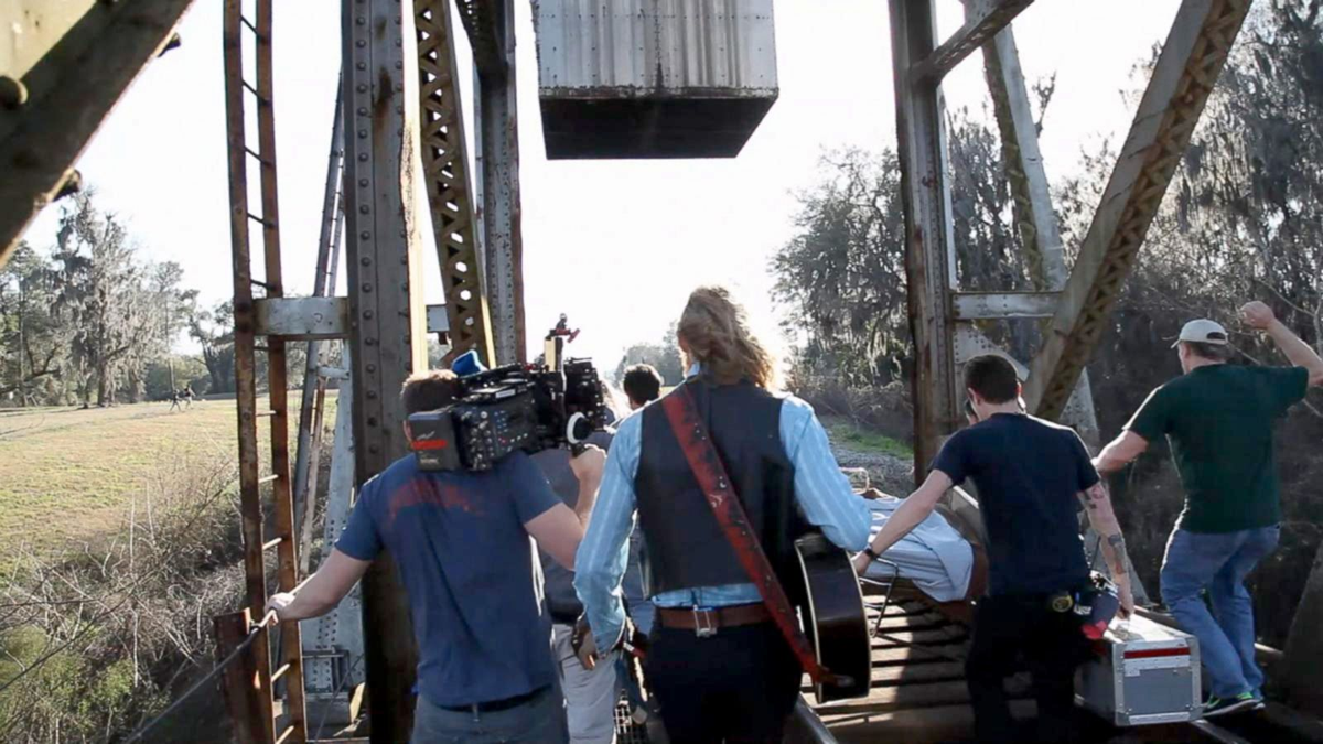 A group of people, one with a camera and one with a guitar, walk across a metal railway bridge. Trees and grass are visible in the background under bright sunlight.