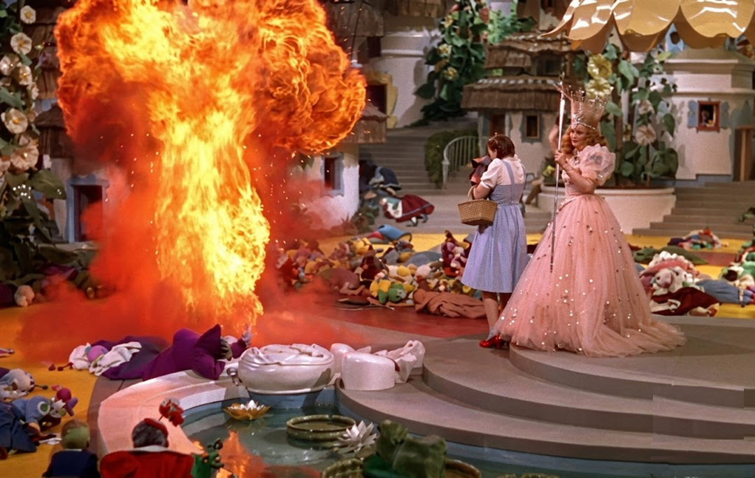Dorothy and Glinda stand together, surrounded by fallen Munchkins, as a massive fiery explosion erupts in the colorful fantasy village scene from The Wizard of Oz.