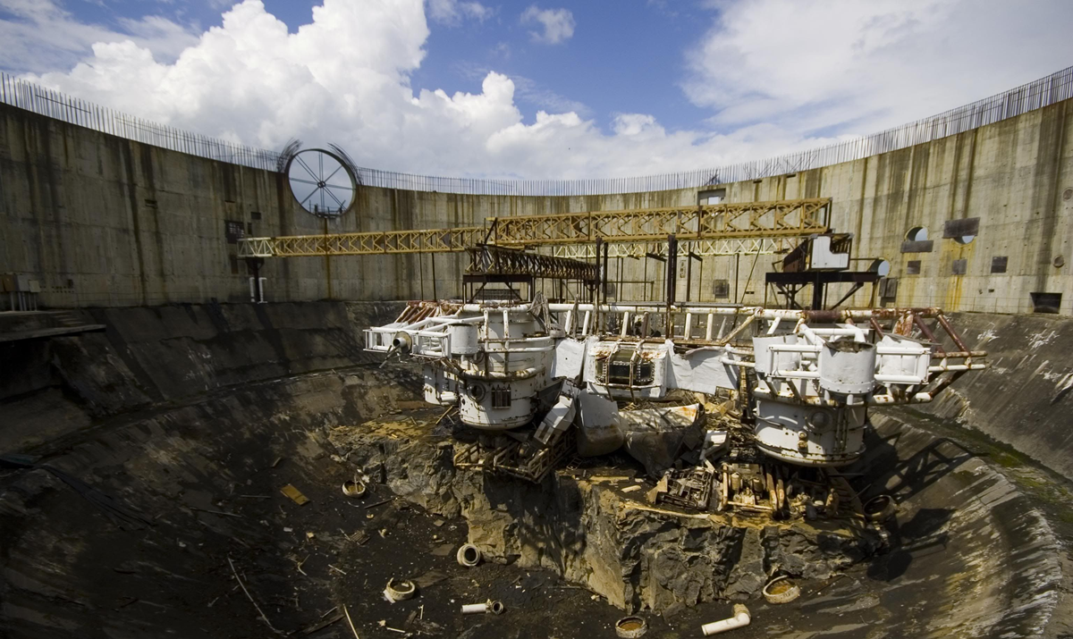 A large, circular, concrete structure with exposed machinery and metal framework stands under a partly cloudy sky. The area appears abandoned and weathered, with debris scattered on the dark ground.