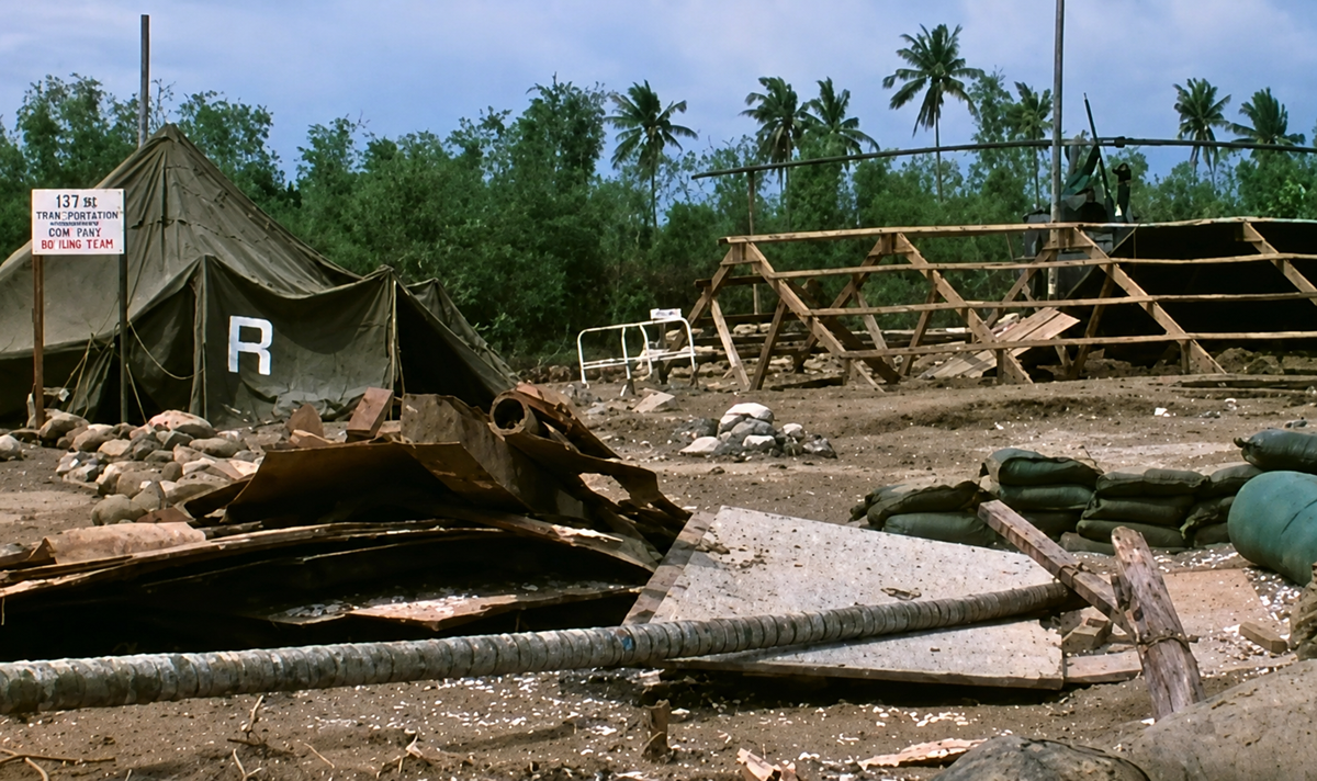 A military camp scene with a tent labeled "R", a helicopter, wooden structures, debris, sandbags, and a sign reading "137 Sht Trans Company" amid palm trees and a clear sky.