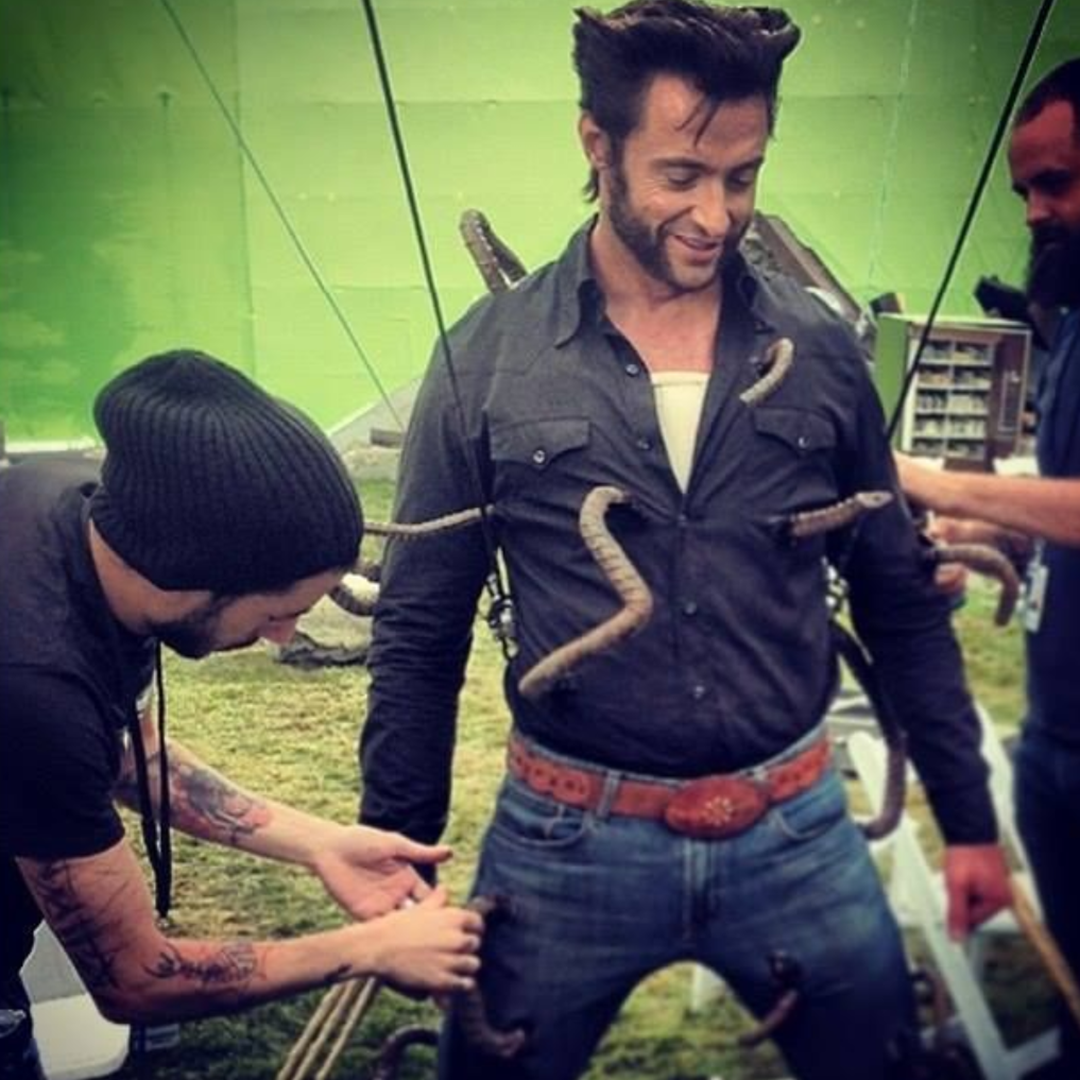 An actor wearing a black shirt and jeans has fake claws attached to his body on a film set, with crew members adjusting props against a green screen background.