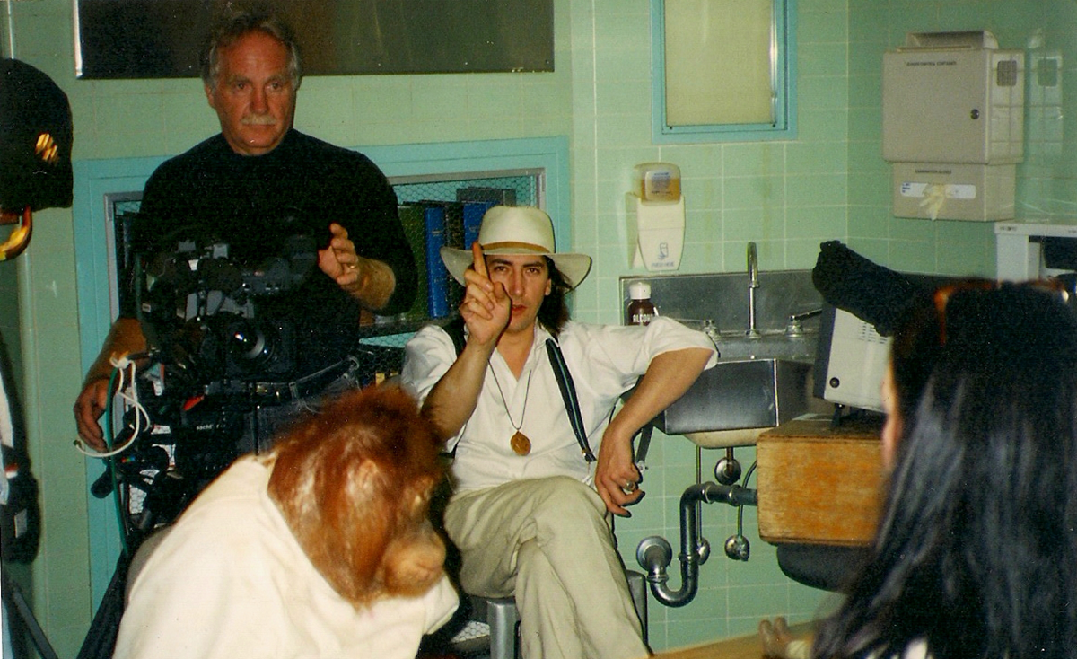 A film set with a camera operator, a man in a white hat gesturing, an orangutan in clothes, and crew members in a room with green tiled walls and medical equipment.
