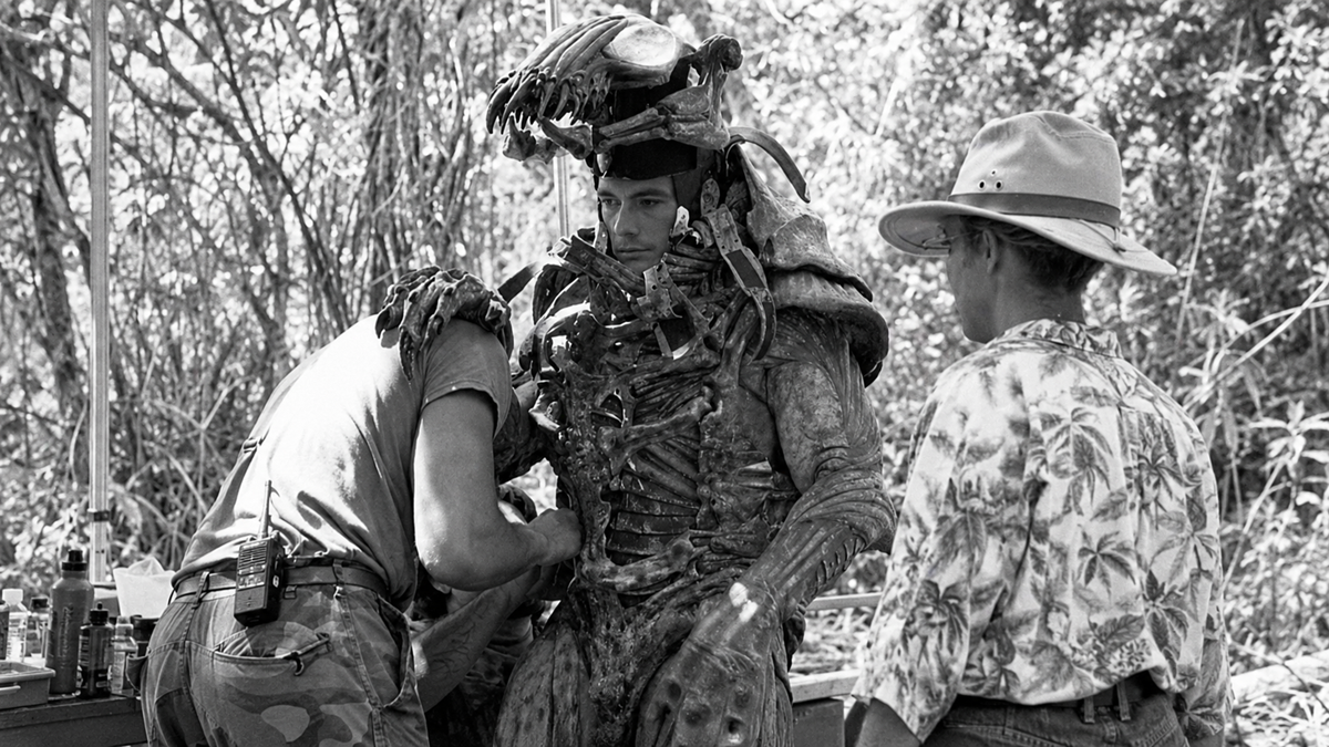 A person wearing a detailed monster costume is being assisted by two crew members outdoors, one adjusting the suit and another watching, surrounded by trees.