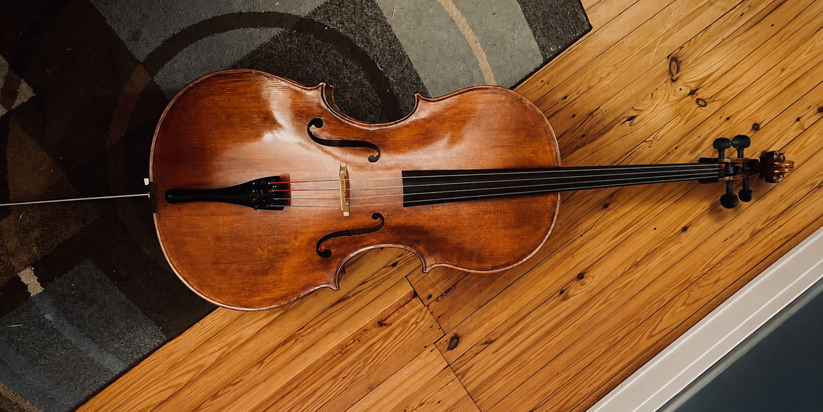 A cello lies on the floor, half on a wooden surface and half on a patterned rug, with its bow resting beside it.