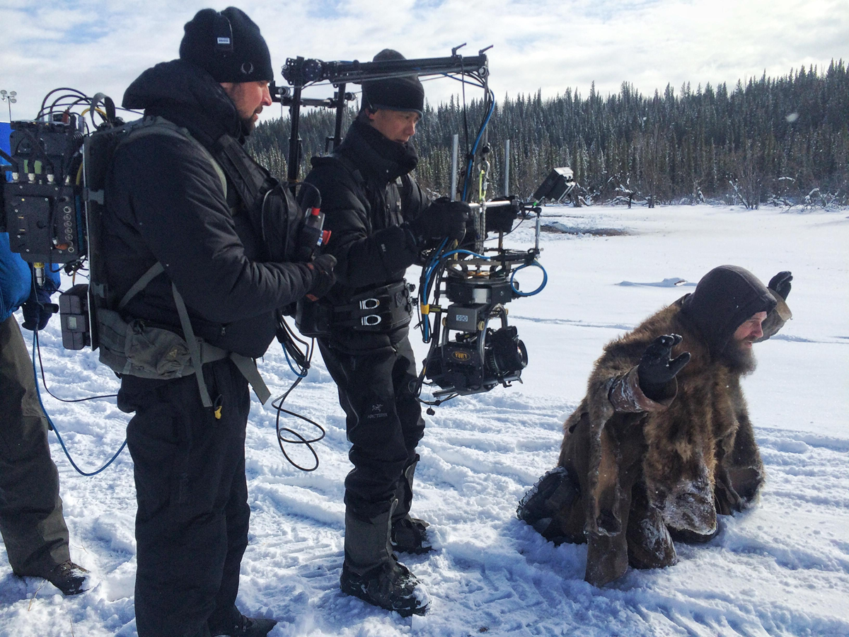 A film crew in winter gear films an actor dressed in fur and kneeling on snow, with snowy trees and a cloudy sky in the background.