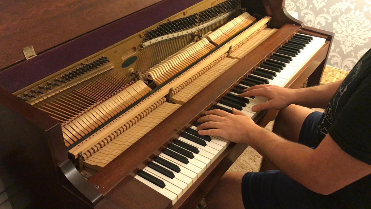 A person plays an upright piano with the front panel removed, revealing the strings and hammers inside; their hands are on the keys as they sit in front of the instrument.