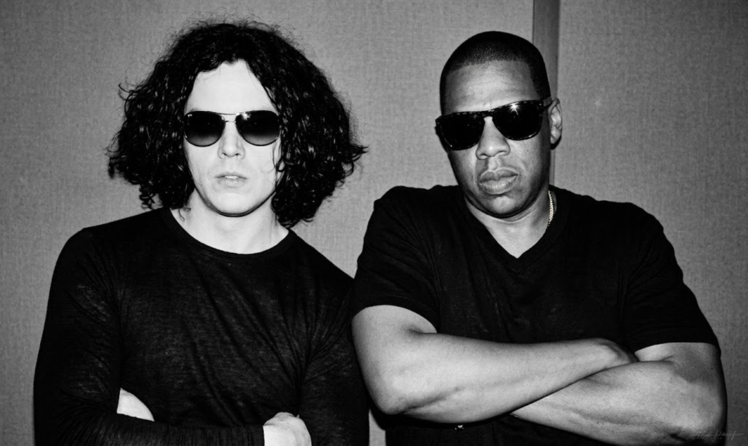 Two men wearing black shirts and sunglasses stand side by side with arms crossed in front of a plain background.