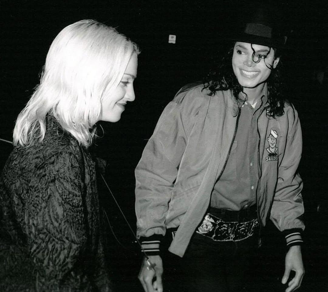 Two people stand together. One person with long dark hair, a hat, and a jacket smiles widely, while the other with light hair looks down, both captured in a candid black and white moment.