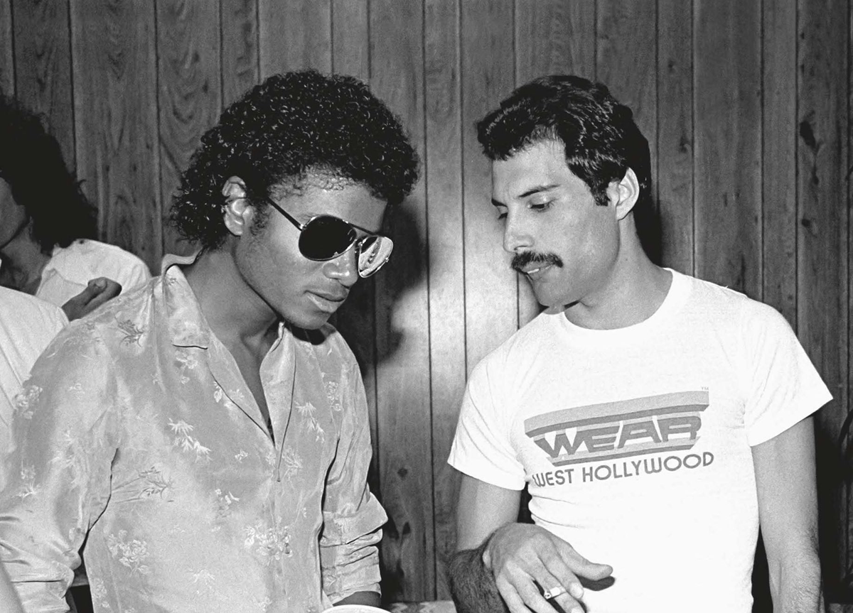 Two men stand together indoors. One wears sunglasses and a patterned shirt, the other has a mustache and a “WEST HOLLYWOOD” t-shirt. They appear to be in conversation, with wood paneling in the background.