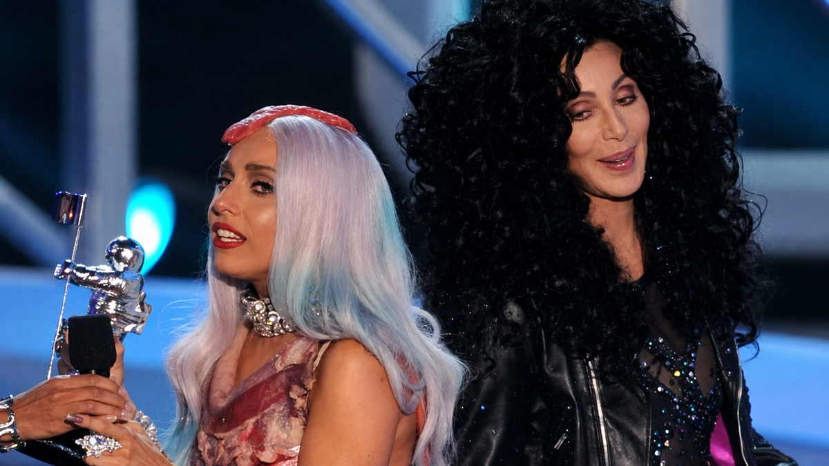 Two women on stage, one with long pastel hair holding a trophy and wearing a dress with a meat-like pattern, the other with long curly black hair in a black outfit, both under stage lights.