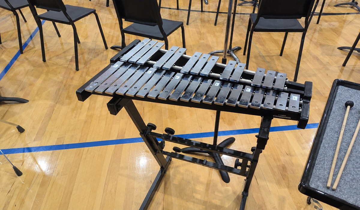 A metal glockenspiel on a black stand is set up on a gymnasium floor, with empty black chairs and music stands in the background. Two mallets rest on a tray beside it.
