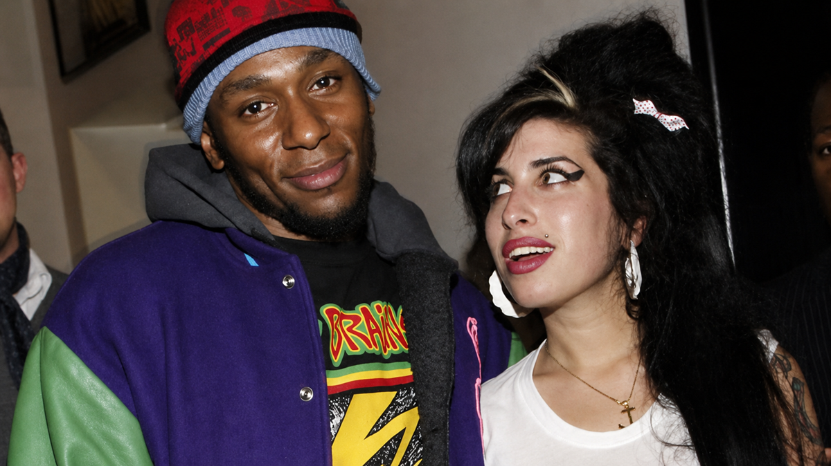 Two people stand close together indoors. The man wears a colorful jacket and blue beanie, smiling slightly. The woman beside him has long black hair, dramatic eyeliner, and large earrings, looking up at him with a surprised expression.