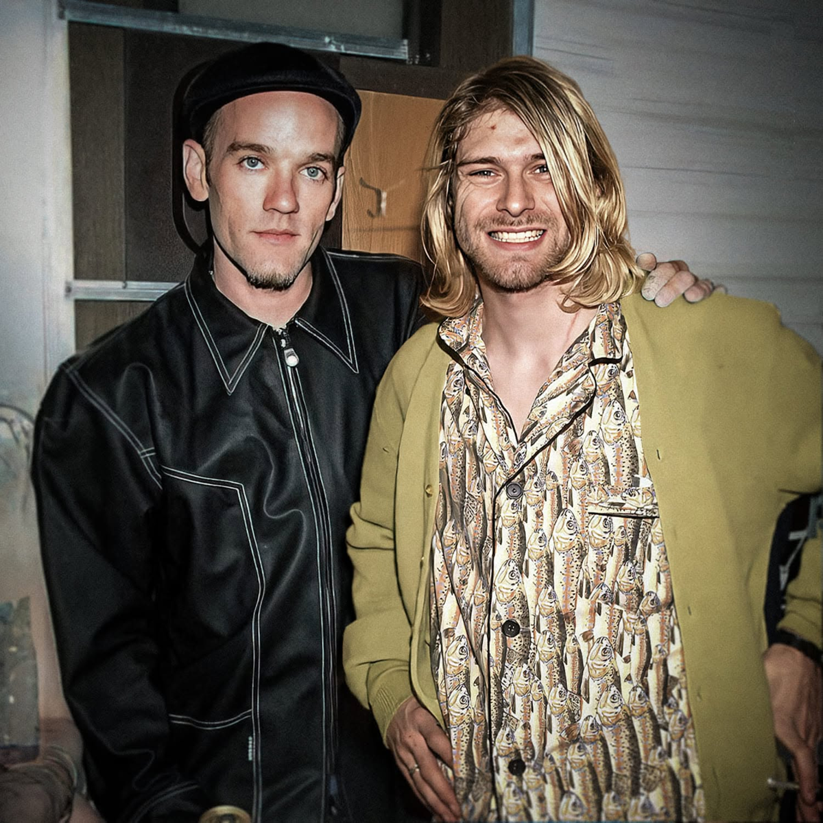 Two men pose together, one wearing a black jacket and flat cap, the other with long blond hair, a patterned shirt, and a green cardigan, smiling with his arm around the other. They stand indoors in front of a wooden door.
