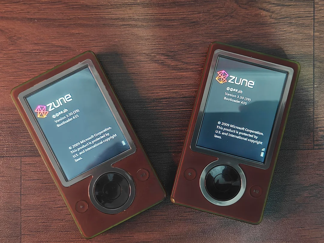 Two brown Microsoft Zune media players sit on a wood surface, both powered on and displaying firmware information screens, showing the Zune logo and device details.