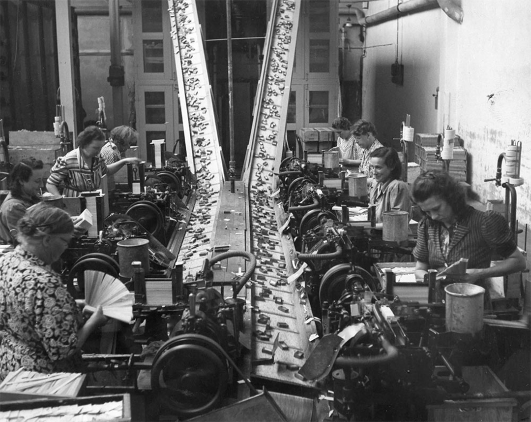 Black-and-white photo of women working at machines in a factory, assembling or packaging items on a conveyor belt. The setting is industrial, with various equipment and workstations visible.