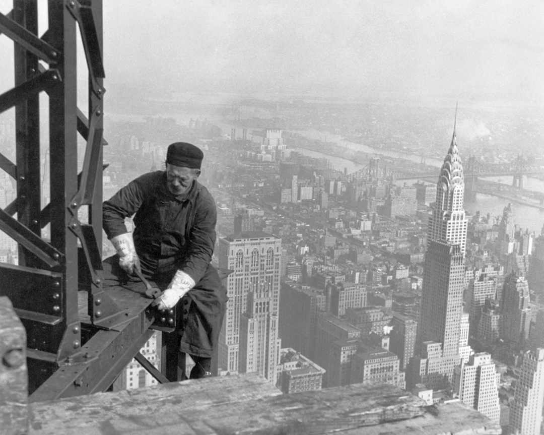 A construction worker in overalls and a cap fastens steel beams high above New York City, with the Chrysler Building and East River visible in the background.