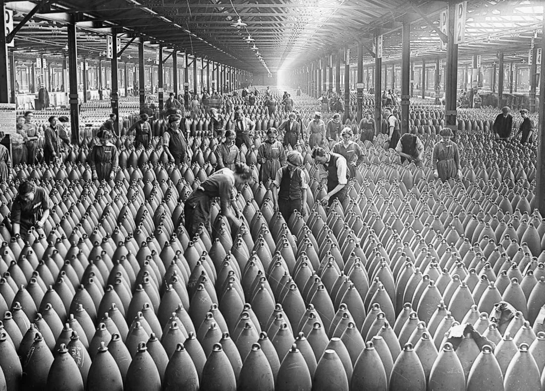 Workers inspect rows of large artillery shells inside a vast, crowded munitions factory, with sunlight streaming through windows and a high beamed ceiling above.