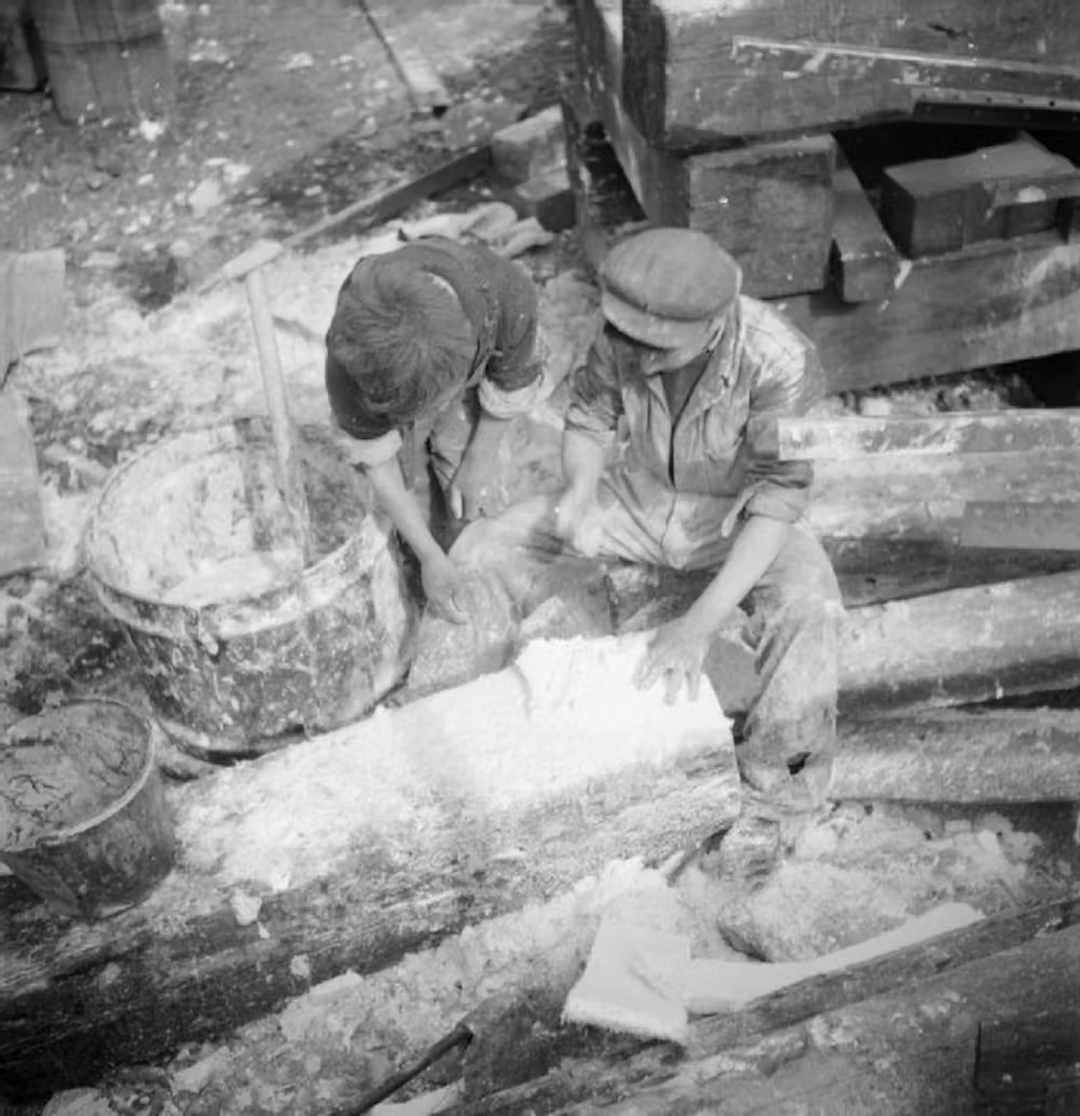 Two workers carve and scrape large wooden beams outdoors, surrounded by sawdust and buckets. One sits while working with a hand tool, both wear work clothes and caps, focused on shaping the timber.