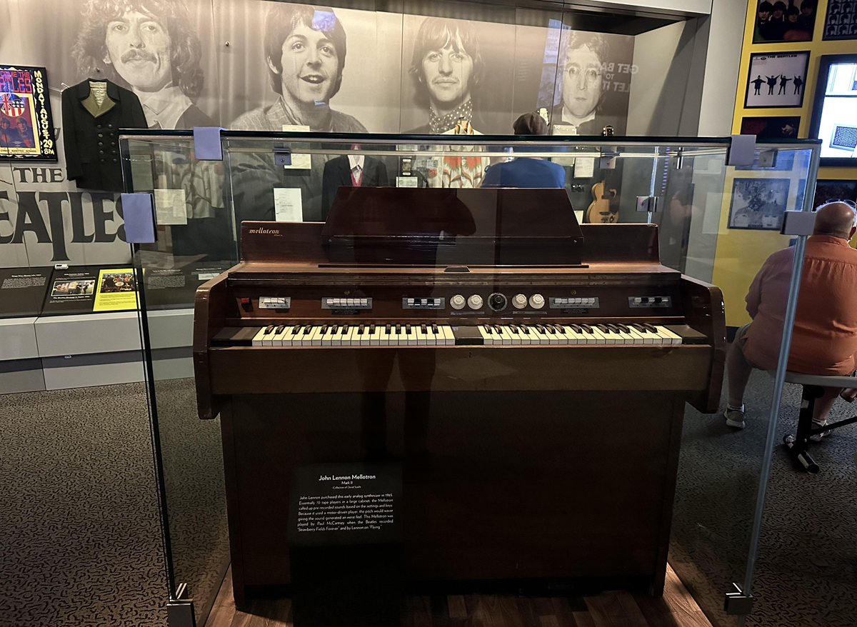 A brown electric organ is displayed in a glass case at a museum. Behind it is a large black-and-white photo of The Beatles and various memorabilia on the walls. A few people are visible in the background.