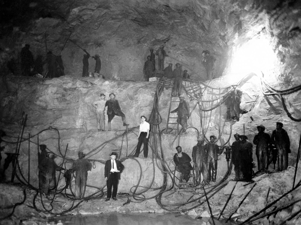 Black-and-white photo of workers in a large cave or mining tunnel, standing among cables and equipment, with light shining from an opening at the upper right.
