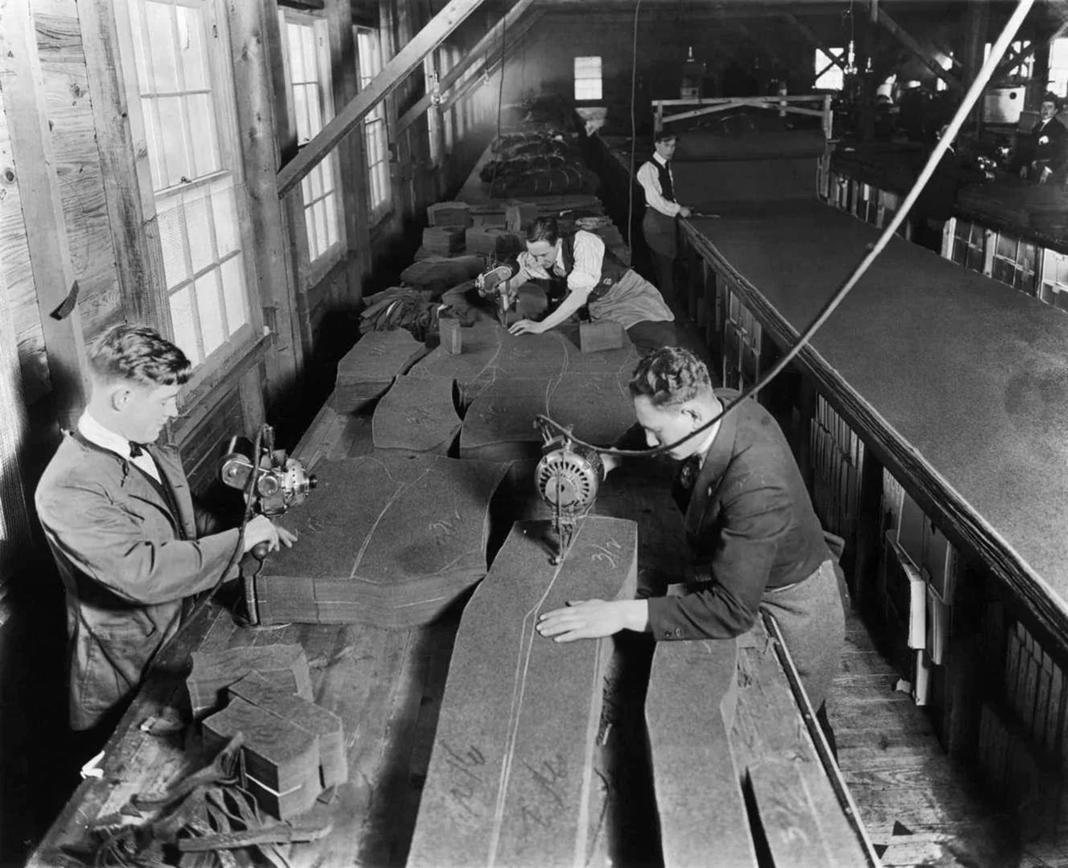Workers using electric cutters shape fabric for men’s suits in a factory, early 20th century. Long tables are covered with layers of fabric, and windows line one side of the workspace.