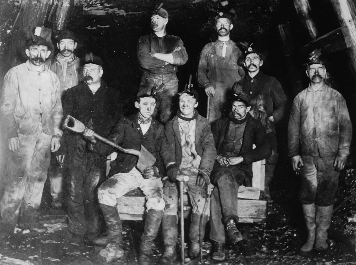 Black and white photo of eleven male coal miners in work clothes and headlamps, sitting and standing inside a dark underground mine, posing for the camera with mining tools.