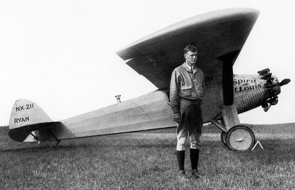 A man stands in front of the Spirit of St. Louis airplane on a grassy field, wearing a flight suit and boots. The aircraft’s name and registration number are visible on its fuselage and tail.