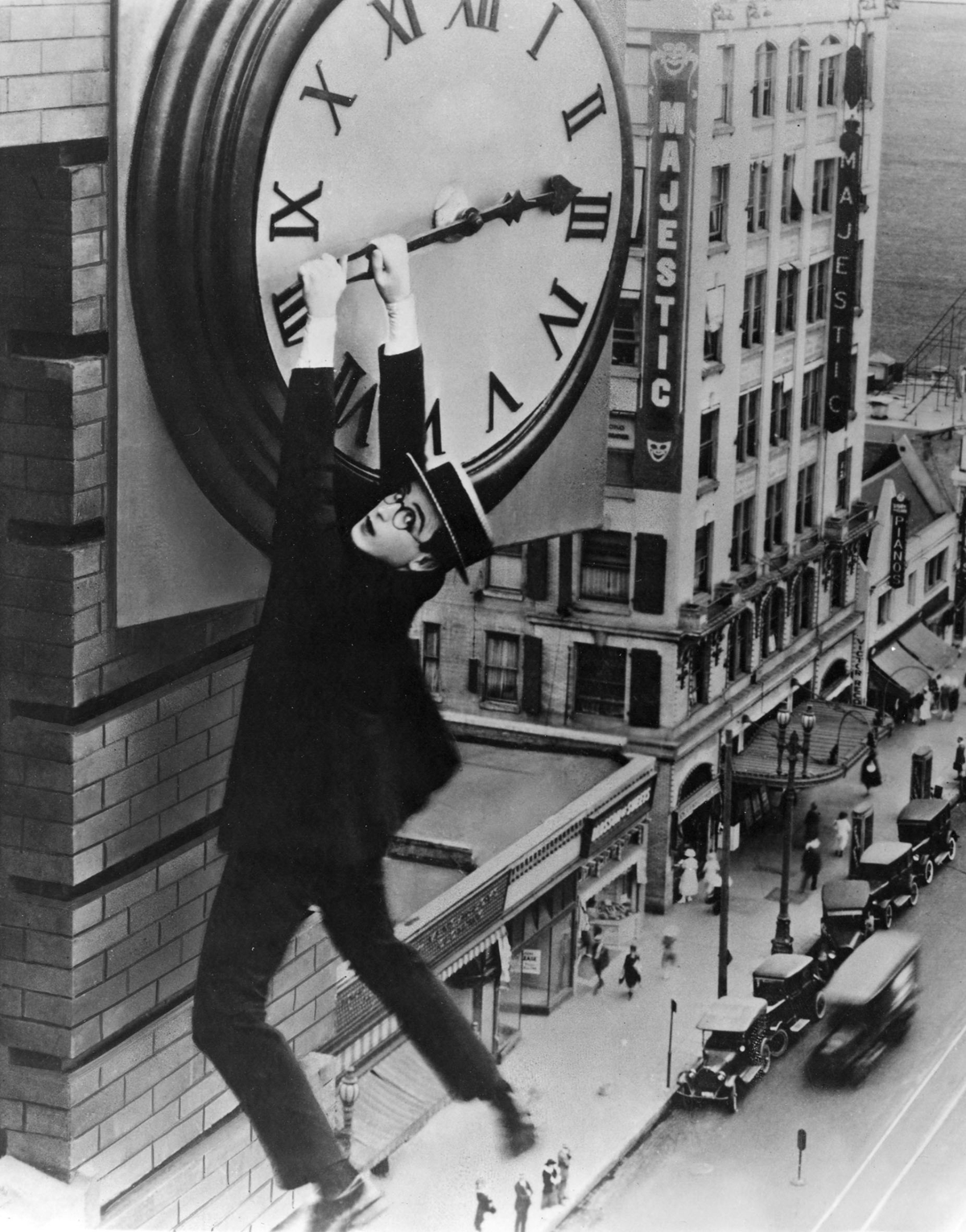A man in a suit and glasses hangs from the hands of a large clock high above a city street, with old cars and buildings below.