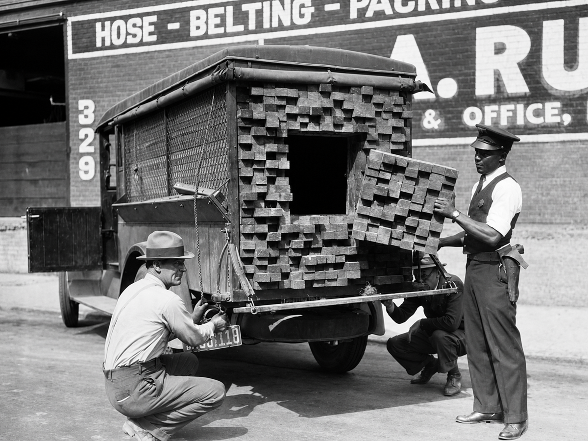 Two men open a van filled with stacked wooden planks, revealing a hidden compartment. A police officer stands beside them. A painted wall advertises hose, belting, and packing services.