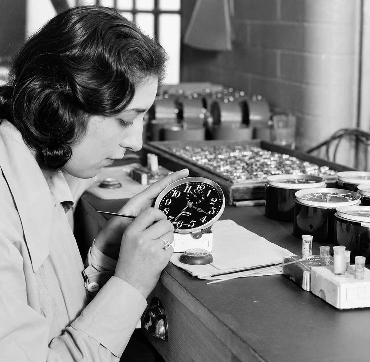 A woman sits at a workbench carefully painting numbers on a clock face, surrounded by clock parts and tools in a workshop setting. Job.