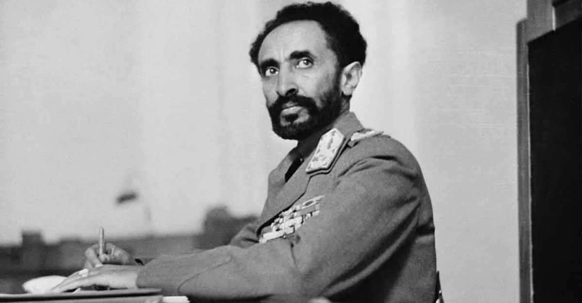 A man in a decorated military uniform sits at a desk, looking upward with a serious expression, holding a pen as if writing or signing a document. Black and white photo.
