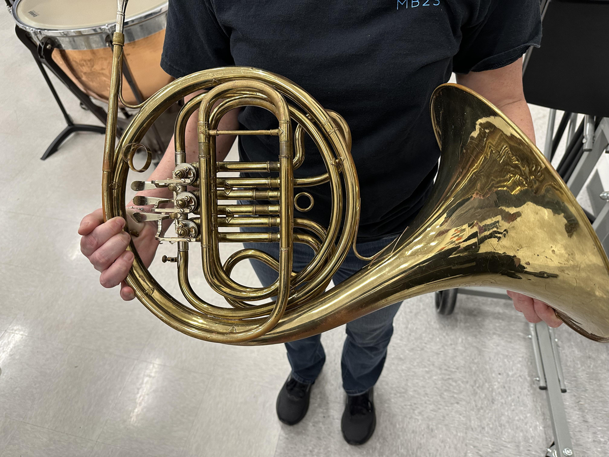 A person wearing a black shirt and jeans holds a brass French horn, standing on a light-colored floor in a music room with drum equipment in the background.