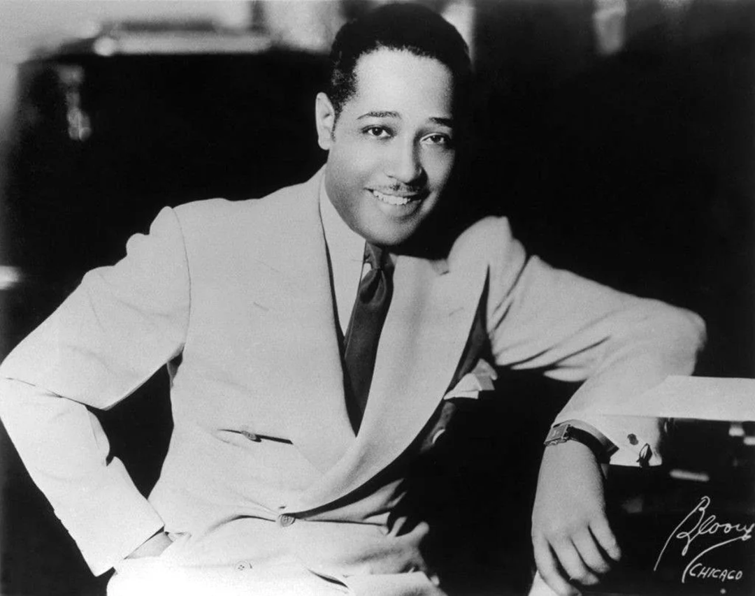 A well-dressed man in a light-colored suit and tie smiles while sitting with one arm resting on a table or piano. The photo is black and white and has a vintage, classic feel.
