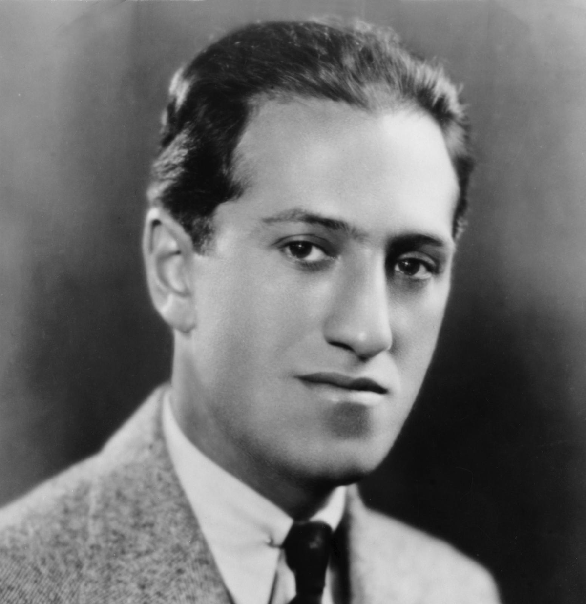 A black-and-white portrait of a young man in a suit and tie, with short dark hair, looking slightly off camera with a neutral expression. The background is plain and softly blurred.