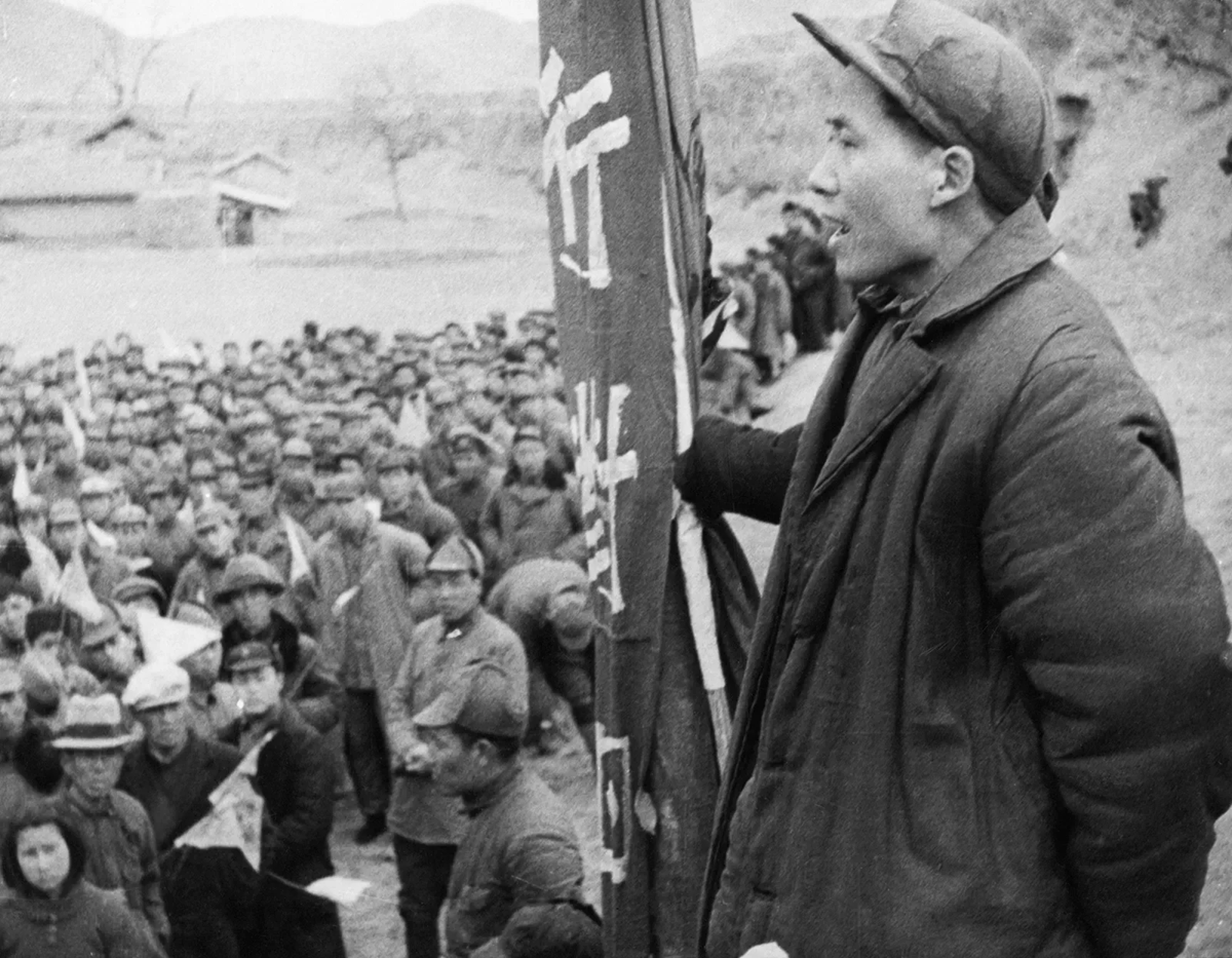 A young person in a cap speaks to a large crowd holding banners with Chinese characters outdoors; people wear winter clothing and hats, and trees and hills are in the background.