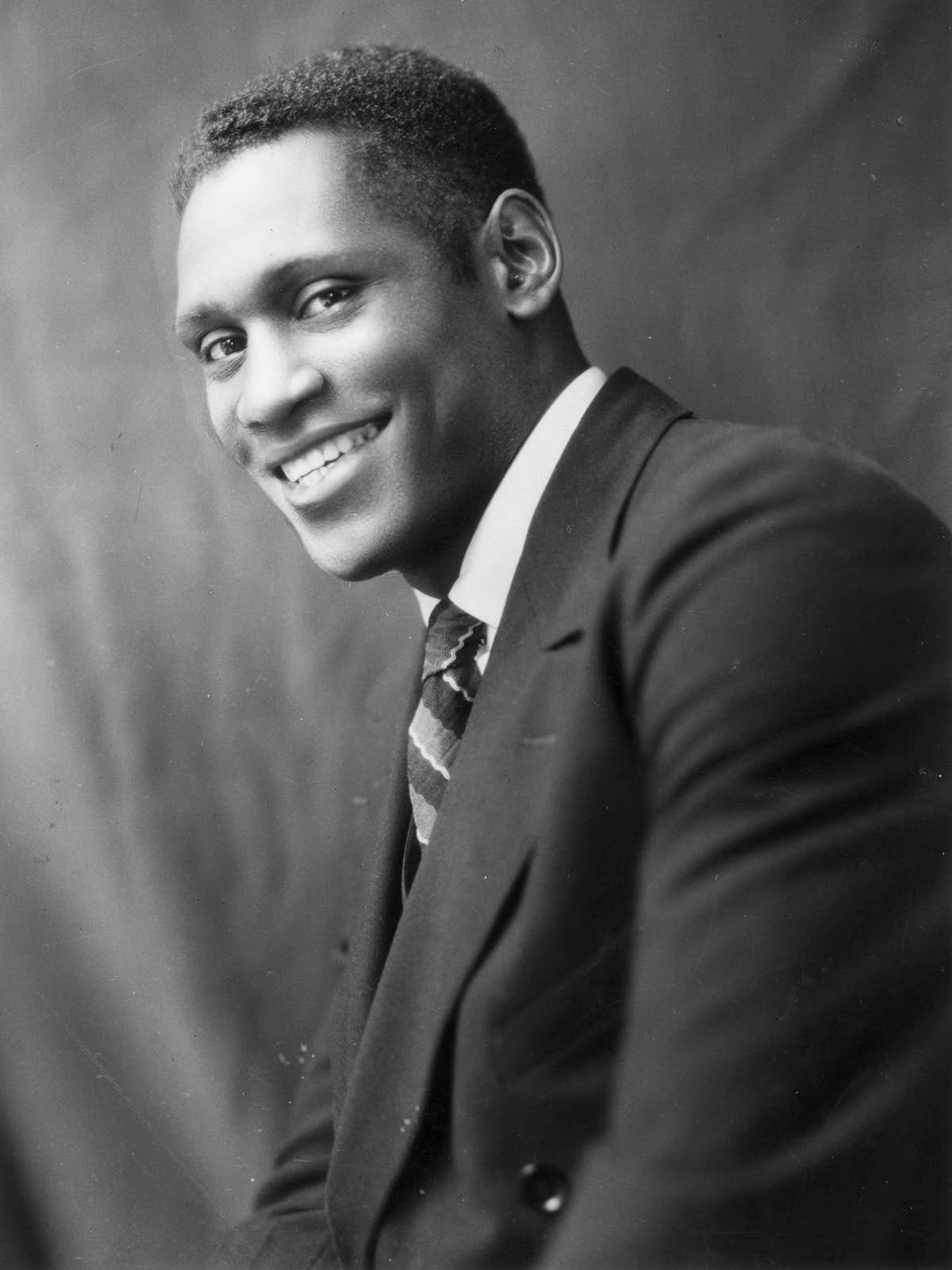 A man in a suit and striped tie smiles at the camera while sitting sideways against a plain, dark backdrop.