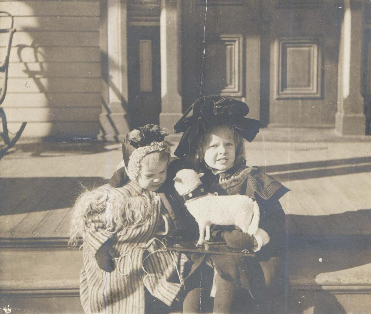 Two young children in old-fashioned winter clothing sit on steps. One holds a doll and the other holds a toy sheep. A building with large doors and a partial sleigh are visible in the background.