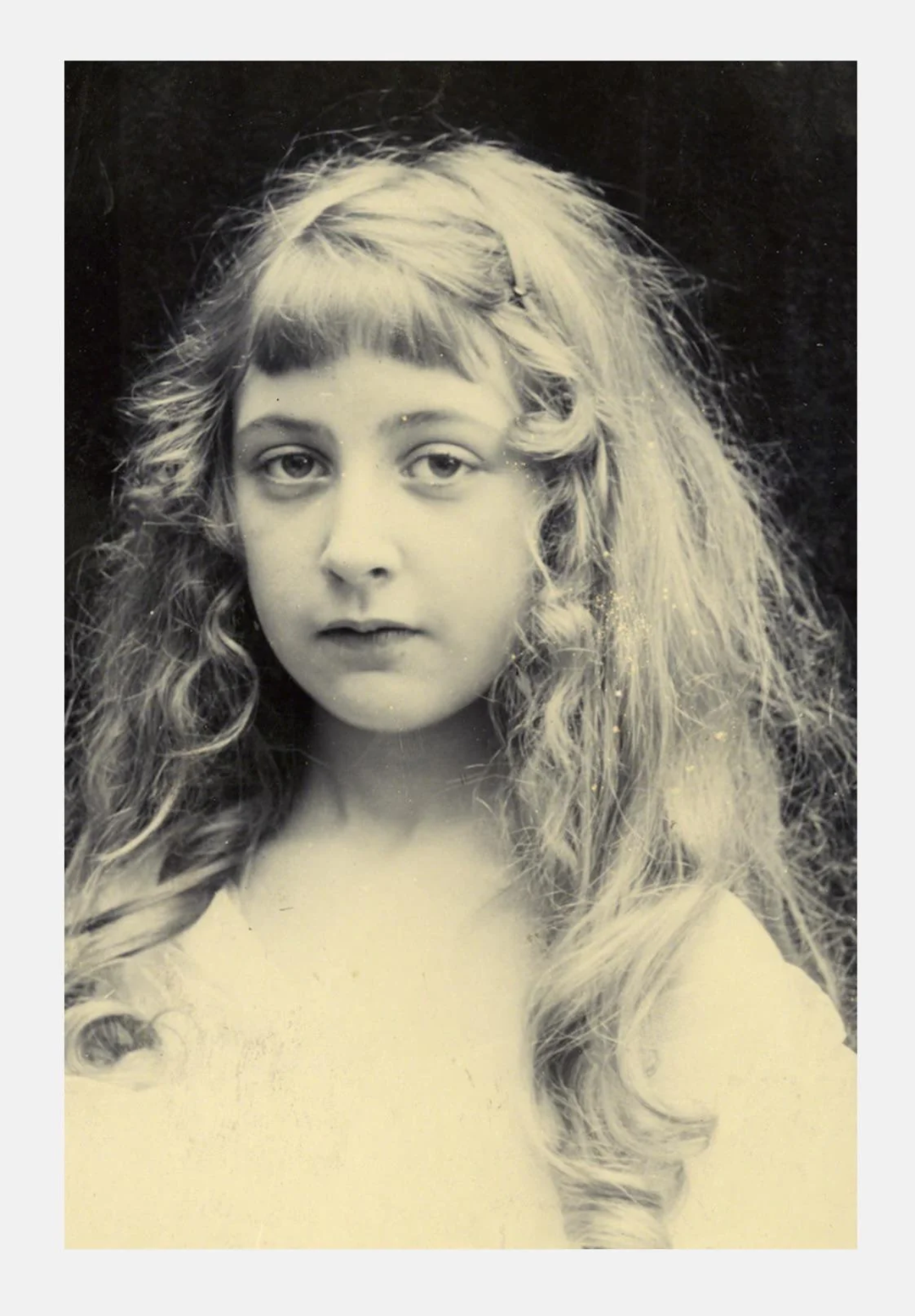 Black-and-white portrait of a young girl with long, wavy hair and bangs, wearing a light-colored top, looking directly at the camera with a neutral expression. 1890s