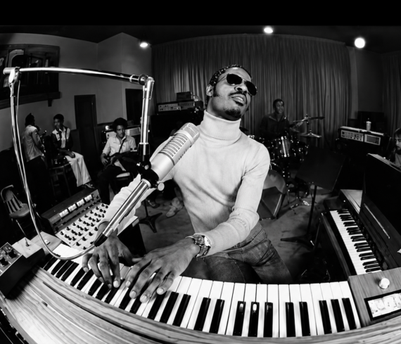 Stevie Wonder wearing sunglasses and a turtleneck plays a keyboard and sings into a microphone in a recording studio, surrounded by a band with drums, guitar, and other instruments.