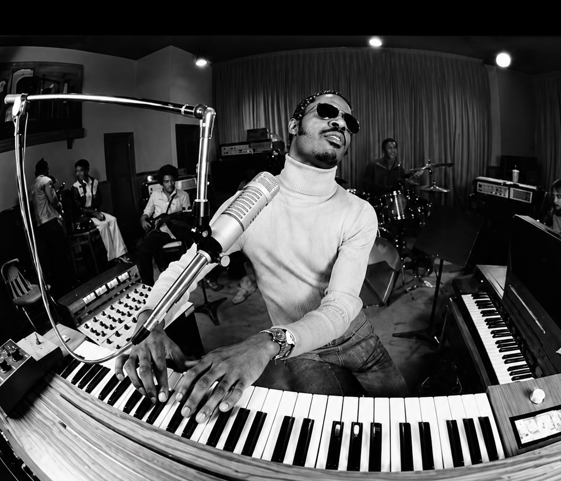 A musician wearing sunglasses and a turtleneck plays a keyboard and sings into a microphone in a recording studio, surrounded by a band with drums, guitar, and other instruments.
