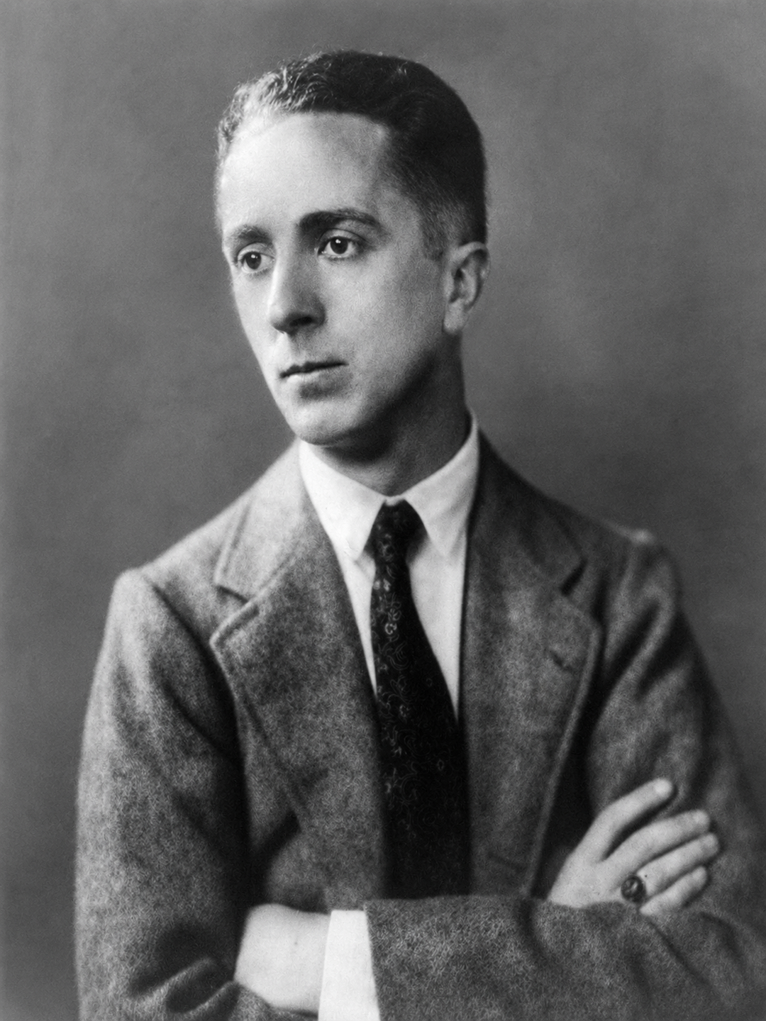 Black-and-white portrait of a young man with short hair, wearing a suit and tie, looking slightly to the side with arms crossed in front of a plain background.