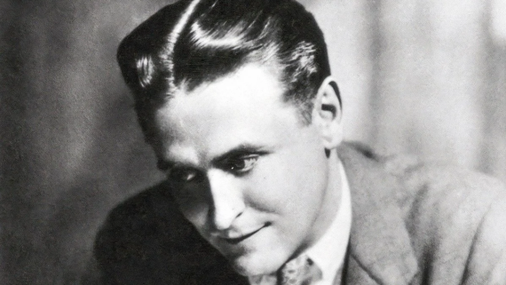 A young man in a suit and tie sits at a desk, looking down and smiling slightly. The image is black and white, and the background is softly blurred.