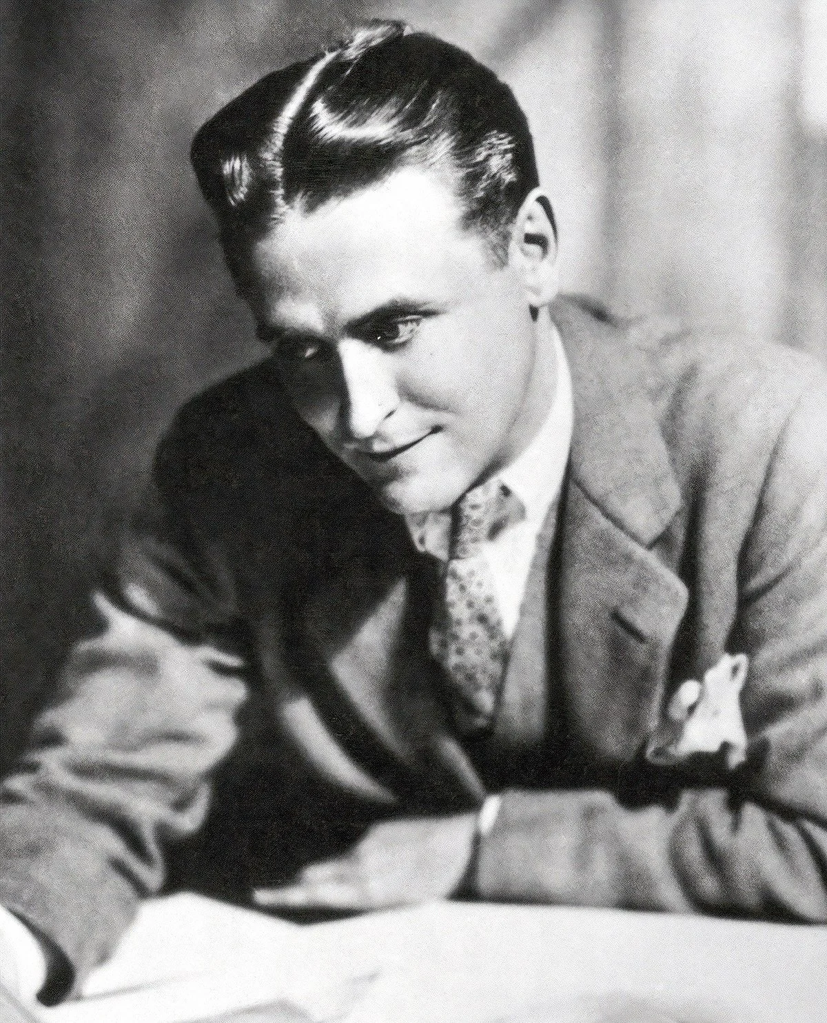 A young man in a suit and tie sits at a desk, looking down and smiling slightly. The image is black and white, and the background is softly blurred.