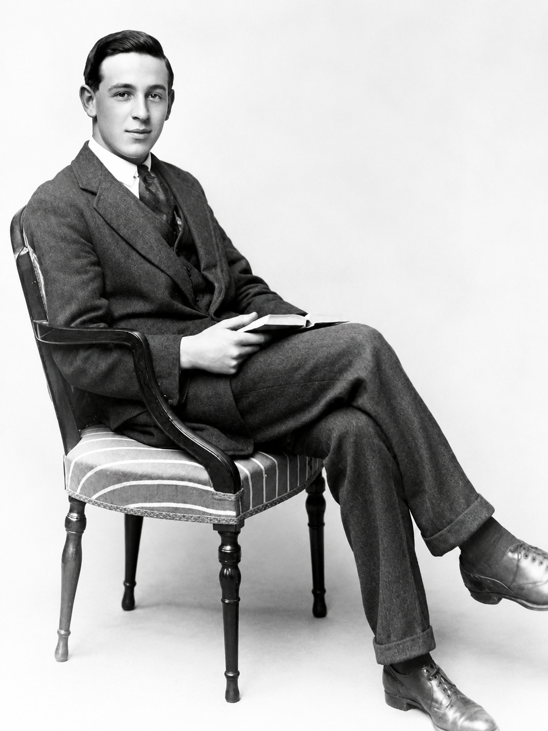 A young man in a suit sits on a striped chair, holding an open book. He looks at the camera with a neutral expression. The photo is black and white.