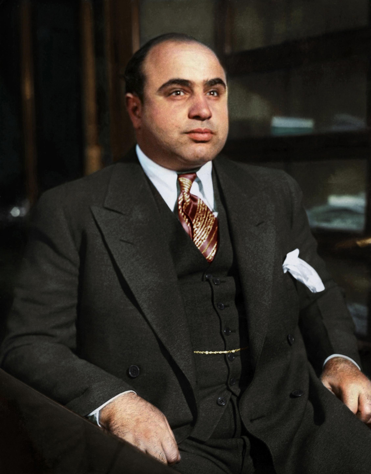 A man in a dark suit, vest, and patterned tie sits at a desk with shelves behind him. He has short dark hair and a white pocket square. The background is blurred.