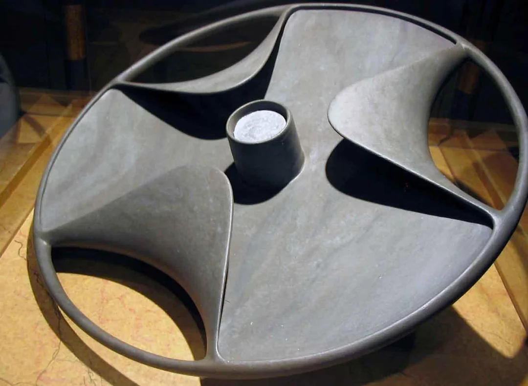 A round, stone disc with curved, wave-like indentations and a raised cylindrical center, displayed on a flat surface under museum lighting.