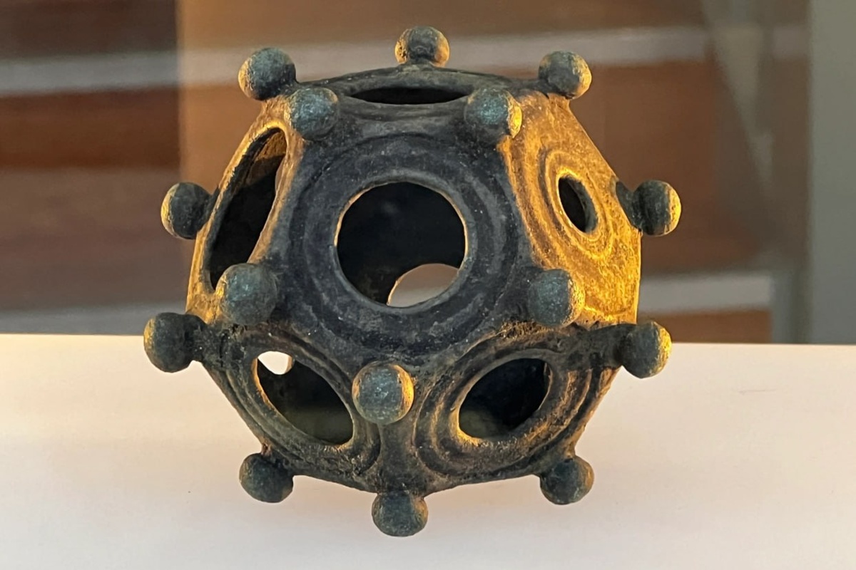 A bronze dodecahedron with circular holes on each face and small knobs at the vertices, displayed on a white surface.