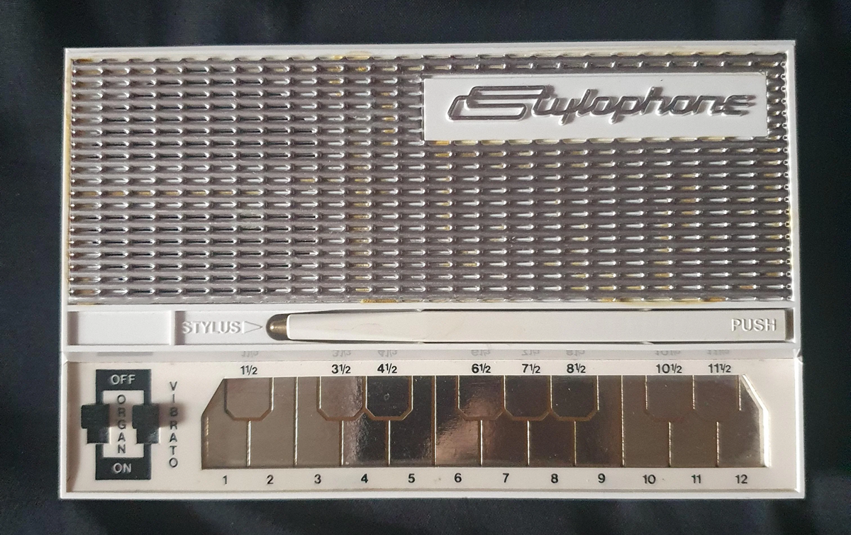 A white and silver Stylophone, a small electronic musical instrument with a metallic keyboard, stylus, and switches labeled "OFF," "ORGAN," and "VIB.ATO." Numbers mark the keys from 1 to 12.